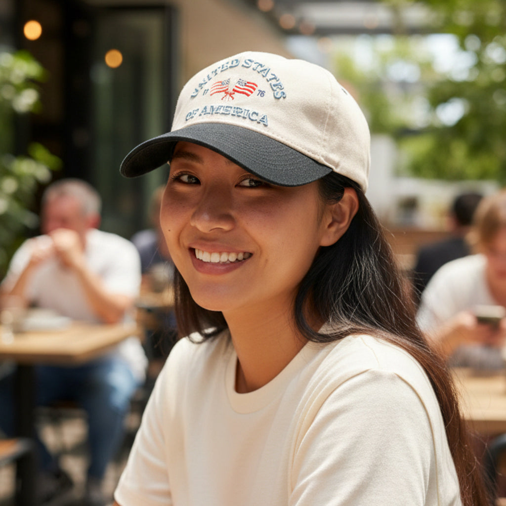 At an outdoor café, a woman in The Darling Effect’s United States of America Classic Canvas Hat and a white shirt smiles, embodying a relaxed vibe while others blur into the background.