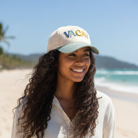 A woman with long blonde hair smiles while tilting The Darling Effect's Vacay Classic Canvas Hat—a white baseball cap with colorful "VACAY" lettering—pairing it effortlessly with a sleeveless white ribbed top against a beige background.