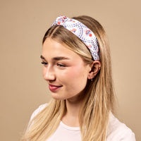 A young woman with long blonde hair smiles softly to the side, wearing The Darling Effect’s All-American Darling Classic Twist Headband in a blue and red pattern, paired with a white t-shirt and small gold hoops against a beige background.