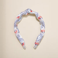 The Darling Effect’s All-American Darling Classic Twist Headband features a charming knot and is decorated with blue stars, red crabs, and small American flags, shown on a plain beige background.
