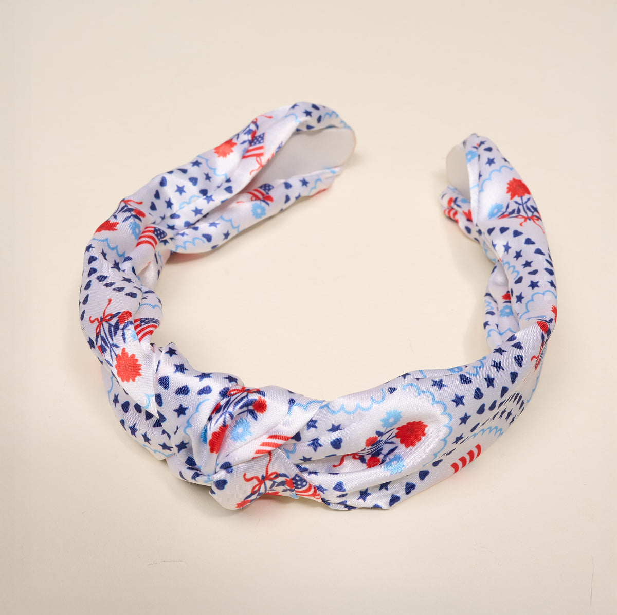 The All-American Darling Classic Twist Headband by The Darling Effect is a versatile accessory featuring a ruched twist design and delicate knot detail. Displayed on a light beige background, it is adorned with red and blue floral and star patterns for a charming touch.