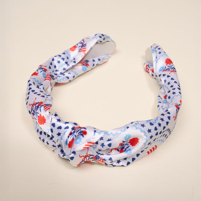 The All-American Darling Classic Twist Headband by The Darling Effect is a versatile accessory featuring a ruched twist design and delicate knot detail. Displayed on a light beige background, it is adorned with red and blue floral and star patterns for a charming touch.