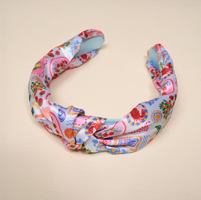 Introducing the Ciao Bella Classic Twist Headband by The Darling Effect—a vibrant silk accessory featuring a twisted design adorned with lively fruit, flower, and decorative item patterns on a soft blue and pink background. Displayed on a simple light surface, this versatile headband adds a delightful touch of charm to any look.