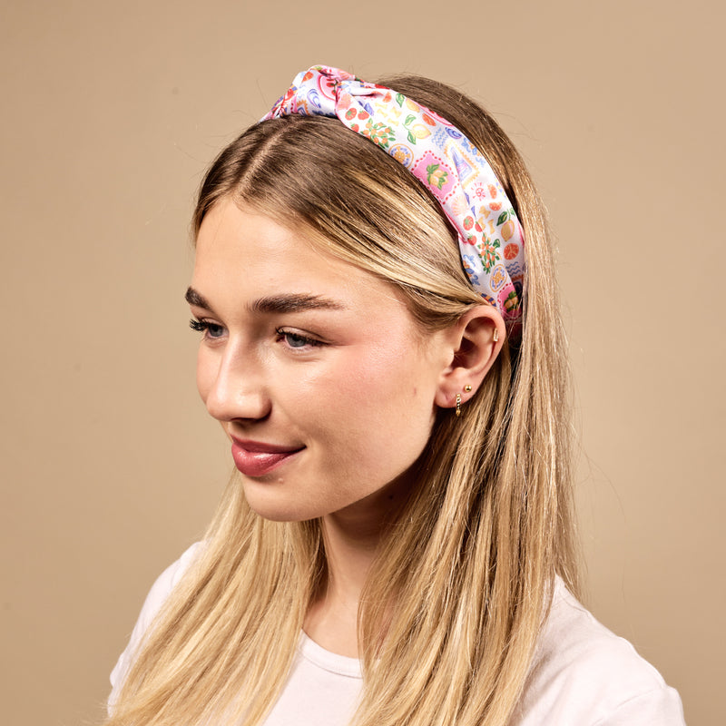 A young woman with long blonde hair wears The Darling Effect's Ciao Bella Classic Twist Headband and a white t-shirt, smiling slightly against a plain beige background.