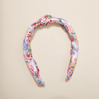 The Darling Effect’s Ciao Bella Classic Twist Headband features a knotted top and vibrant floral-paisley print in red, blue, green, and yellow on white—a stylish, versatile accessory displayed on a beige surface.