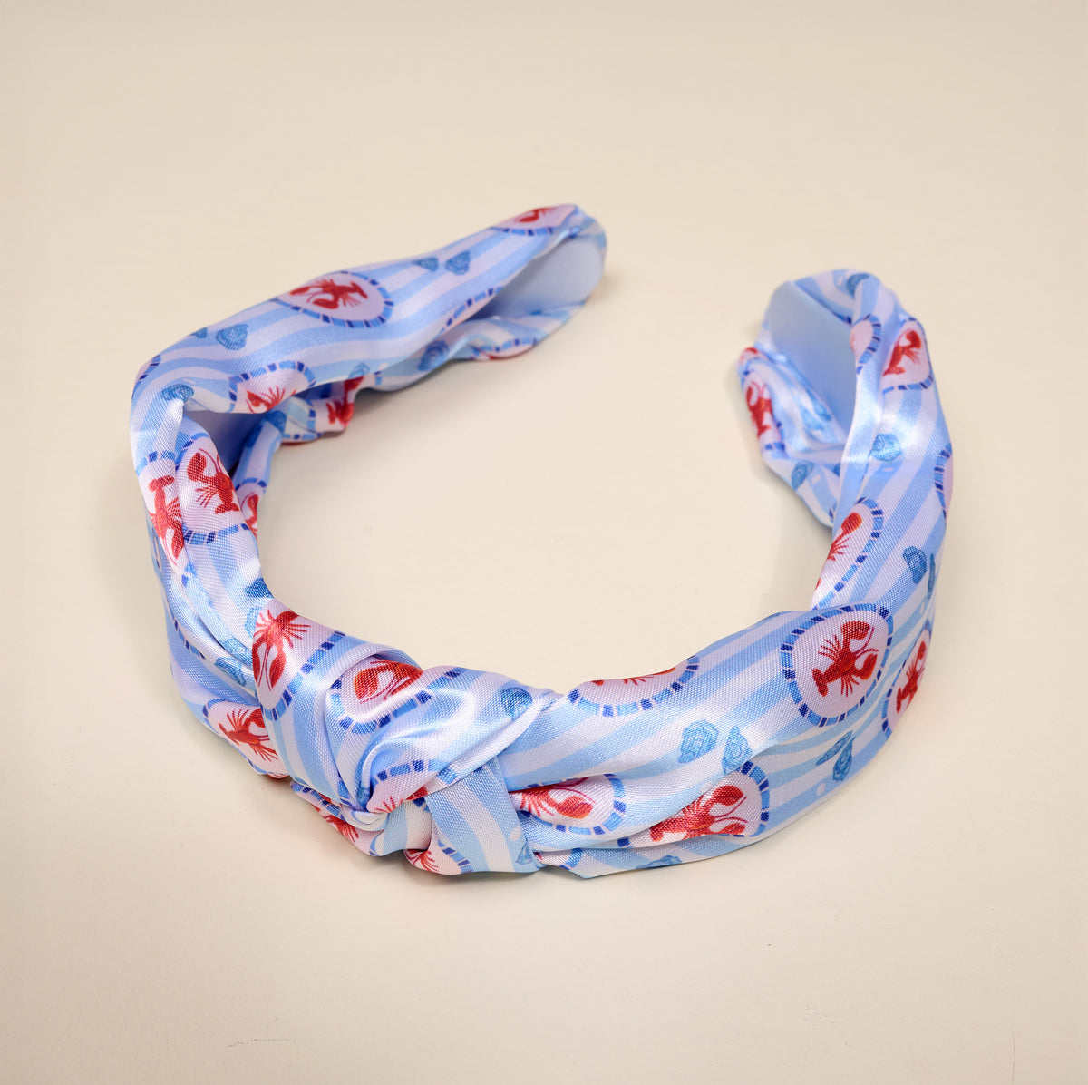 The Darling Effect’s Harbor House Classic Twist Headband features blue satin with white stripes and whimsical red lobster prints, elegantly knotted at the front and displayed on a beige background—this versatile accessory brings playful charm to any outfit.