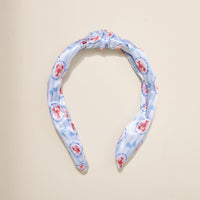 The Darling Effect’s Harbor House Classic Twist Headband features a light blue fabric with red lobsters and blue seashells, styled in an elegant top knot and shown on a beige background—a playful coastal accessory for any outfit.