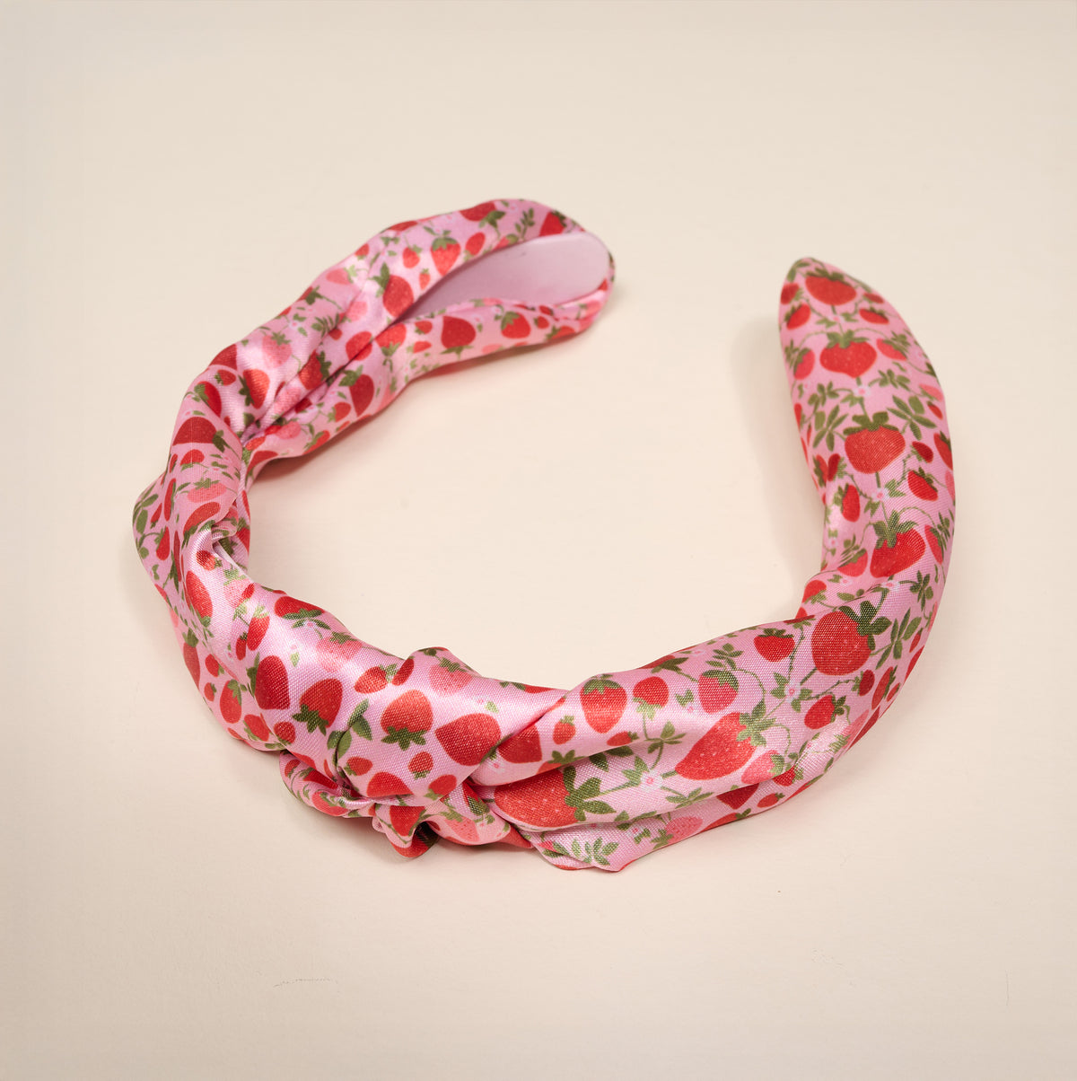 The Strawberry Fields Classic Twist Headband by The Darling Effect is a pink satin accessory adorned with a charming strawberry pattern featuring red strawberries and green leaves, shown against a simple light background. Both stylish and functional, this versatile headband effortlessly adds a playful yet chic touch to any outfit.