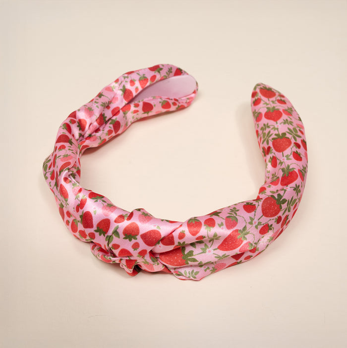 The Strawberry Fields Classic Twist Headband by The Darling Effect is a pink satin accessory adorned with a charming strawberry pattern featuring red strawberries and green leaves, shown against a simple light background. Both stylish and functional, this versatile headband effortlessly adds a playful yet chic touch to any outfit.