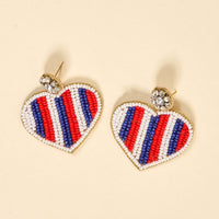 Americana Heart Beaded Earrings by The Darling Effect feature vibrant heart shapes with red, white, and blue bead stripes and a clear rhinestone cluster. These playful earrings are shown on a light beige background.