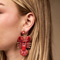 A close-up of a woman’s ear wearing The Darling Effect Lobster Beaded Earrings highlights her love for vibrant accessories. With straight, light brown hair and fair skin, she showcases the playful charm of these large, red, lobster-shaped hand-beaded earrings.