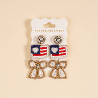The Darling Effect’s Star Spangled Bow Beaded Earrings feature hand-beaded American flag hearts, accented with dangling white and gold bows and sparkling rhinestones, presented on a branded card.