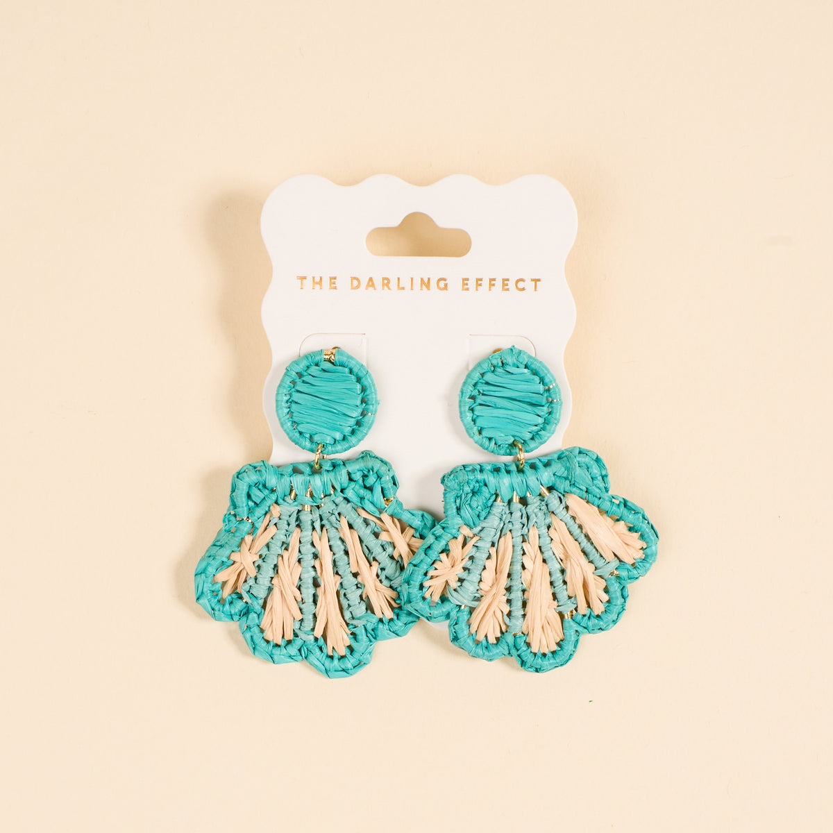 The Darling Effect's Aqua Cabana Breeze Earrings—turquoise and beige woven shells on a white card with the brand print—are lightweight, boho-chic accessories perfect for everyday style against a light beige backdrop.
