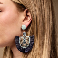 A woman wears The Darling Effect's Navy Fringe Earrings—boho style with a light blue woven stud, navy raffia fan-shaped fringe, and gold bead accents. These are perfect lightweight earrings for everyday wear.