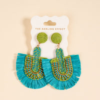 The Darling Effect's Teal Fringe Earrings feature round green tops, blue fringe, and green, blue, and gold beadwork. Lightweight and perfect for everyday wear, they come displayed on a branded card.