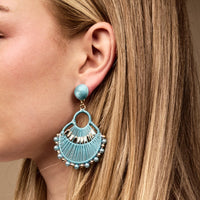 A woman's ear with blonde hair showcases The Darling Effect's Teal Paloma Drop Earrings—boho, fan-shaped with gold accents and beads. These lightweight earrings are perfect for adding stylish flair to everyday looks.