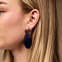 A close-up of a woman wearing The Darling Effect's Navy Pretty and Polished Earrings—statement drops featuring gold ball studs and cascading navy velvet petal charms, perfect for everyday wear, shown against her blonde highlighted hair.