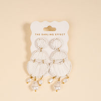 The Darling Effect's White Pearl Affair Earrings feature white statement design with gold accents and pearl-like beads. Lightweight for daily wear, they're displayed on a card against a light beige background.