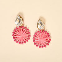 The Darling Effect's Hot Pink Radiant Gem Earrings are lightweight, boho-style earrings with clear teardrop gems atop round pink woven designs accented by small pink beads. Set against a light beige background.