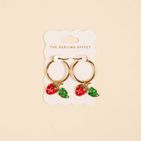The Berrylicious Signature Hoop Earrings by The Darling Effect feature gold hoops adorned with vibrant red strawberry and green leaf charms. Displayed on a cream card labeled with the brand name against a beige background, these lightweight earrings add a playful touch to any look.