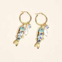 The Darling Effect's Catch of the Day Signature Hoop Earrings feature gold hoops with dangling koi fish charms, blue and purple enamel accents, faux pearls, and clear gemstones—perfect for adding playful flair to your everyday style. Displayed on a light background.