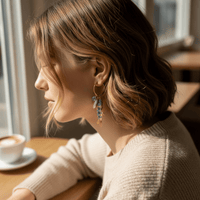 A woman with short, wavy hair sits by a café window, wearing a beige sweater and The Darling Effect's Catch of the Day Signature Hoop Earrings. Sunlight highlights her thoughtful gaze as she enjoys coffee, embodying effortless style.