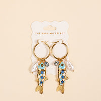 The Darling Effect's Catch of the Day Signature Hoop Earrings—gold hoops with a blue and gold fish charm, white pearl, and round gem—add playful elegance to everyday looks. Shown on a branded card against a beige background.