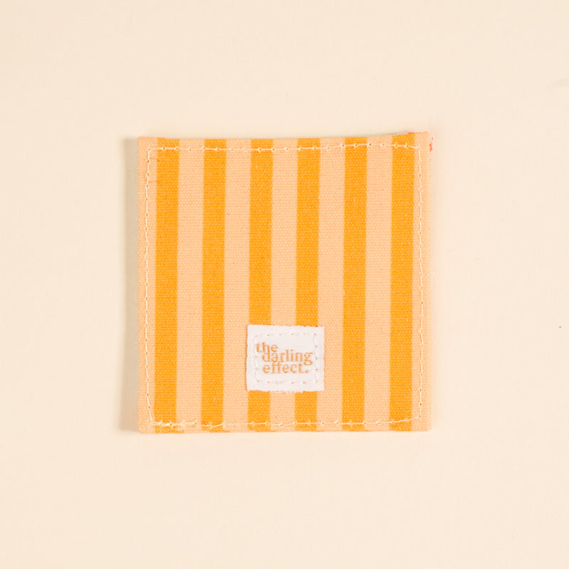 A square fabric coaster with vertical yellow and light beige stripes, white stitching, and a small "The Darling Effect" label. Perfect paired with the Italian Summer Corner Keeper Fabric Bookmark or as a thoughtful gift for book lovers.