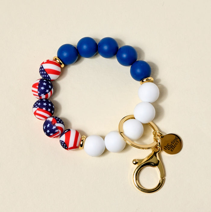 The Darling Effect Americana Hands-Free Keychain Wristlet features blue, white, and flag-patterned silicone beads, a gold clasp, and a round engraved charm—a stylish everyday hands-free key holder.
