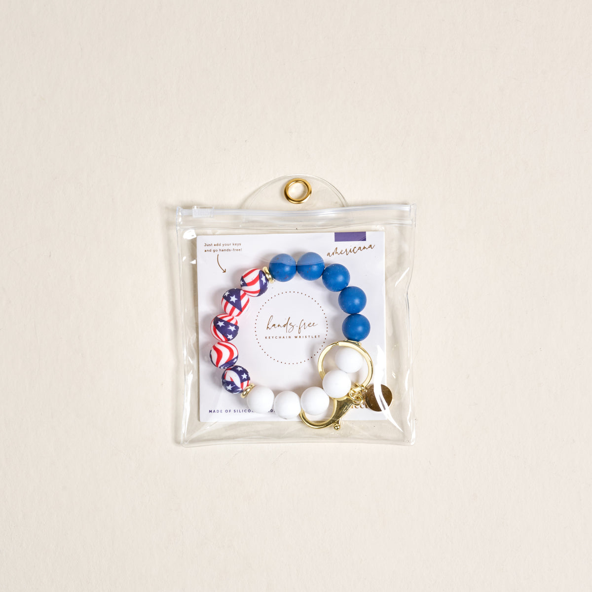 The Darling Effect Americana Hands-Free Keychain Wristlet features blue, white, and red round beads in patriotic patterns on a gold keyring with clasp, packaged in a clear zippered pouch against a neutral background.