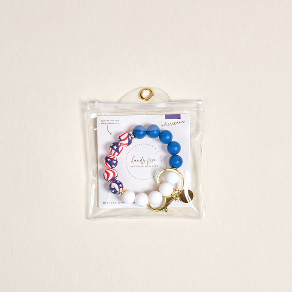 The Darling Effect Americana Hands-Free Keychain Wristlet features blue, white, and red round beads in patriotic patterns on a gold keyring with clasp, packaged in a clear zippered pouch against a neutral background.
