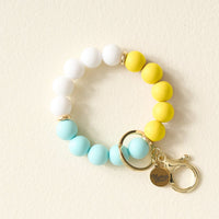 The Darling Effect's Ray of Sunshine Hands-Free Keychain Wristlet features white, yellow, and light blue silicone beads, gold key rings, and a charm engraved with The Giving Effect—perfect for holding keys stylishly on the go.