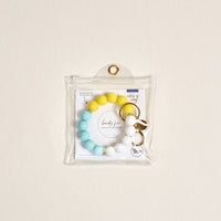 The Darling Effect's Ray of Sunshine Hands-Free Keychain Wristlet, featuring yellow, blue, and white beads, is displayed in a clear plastic pouch with a gold grommet against a light beige background.