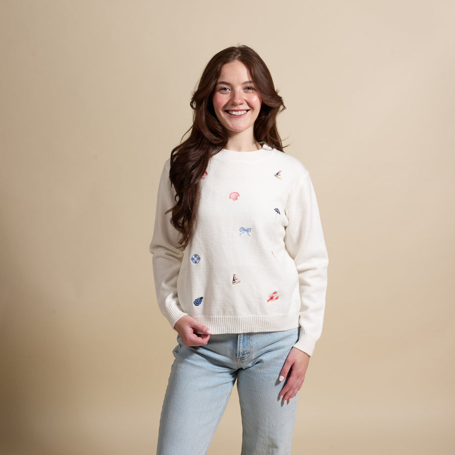 A smiling woman with long brown hair wears a cream sweater with small colorful embroidered designs and light blue jeans, standing against a beige background.