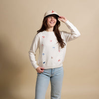 A smiling woman wears The Darling Effect’s Seaside Haven Refined Knit Sweater—an elegant piece with colorful embroidery—paired with light blue jeans and a beige cap, standing against a plain beige background.