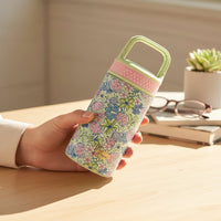 A hand holds The Darling Effect’s Coastal Cove 6 oz Lil' Bitty Bottle, adorned with floral and sea turtle patterns, a pink band, and light green handle—making it the perfect compact travel companion for hydration on the go.