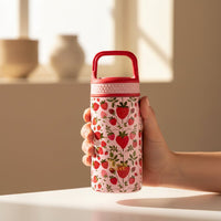 A hand holds The Darling Effect’s Strawberry Fields 6 oz Lil’ Bitty Bottle—pink, adorned with red strawberries and green leaves, plus a red handle—on a white surface. Blurred vases and sunlight in the background complete this stylish on-the-go hydration.