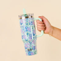 A hand holds the-darling-effect Coastal Cove 40 oz Lifestyle Flip Straw Tumbler, featuring a tropical print with pink flowers and blue sea turtles, a leak-resistant design, mint green handle, and easy-sip flip straw.