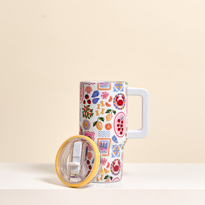 The Darling Effect Ciao Bella 20 oz Lifestyle Flip Straw Tumbler features a vibrant fruit and geometric pattern. It stands upright on a white surface, with its clear lid featuring a yellow rim beside it, set against a light beige background.