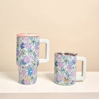 Two Coastal Cove 20 oz Lifestyle Flip Straw Tumblers by The Darling Effect, featuring pink, blue, and green floral patterns and large white handles, rest on a white surface. Each insulated stainless steel tumbler includes a pink or clear lid.