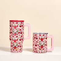 Two pink mugs with handles from The Darling Effect—one a tall Strawberry Fields 20 oz Lifestyle Flip Straw Tumbler (insulated stainless steel with lid), the other shorter and lidless—display strawberries and hearts on a white surface against a beige background.