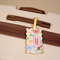 A close-up of a beige suitcase with brown accents, featuring The Darling Effect's Ciao Bella Jetsetter Luggage Tag—a decorative tag with colorful illustrations, gold trim, and a privacy flap—attached to the handle.