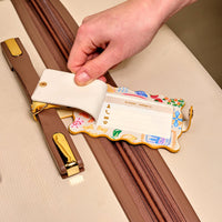A hand lifts the privacy flap of The Darling Effect’s Ciao Bella Jetsetter Luggage Tag, a colorful travel accessory on a beige suitcase with brown and gold details, revealing a blank address label underneath.