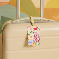 The Darling Effect’s Ciao Bella Jetsetter Luggage Tag, in cream with a playful crab, tomatoes, fruit slices, and sardines design, hangs from a beige suitcase handle—adding charm and keeping your info secure with its privacy flap.
