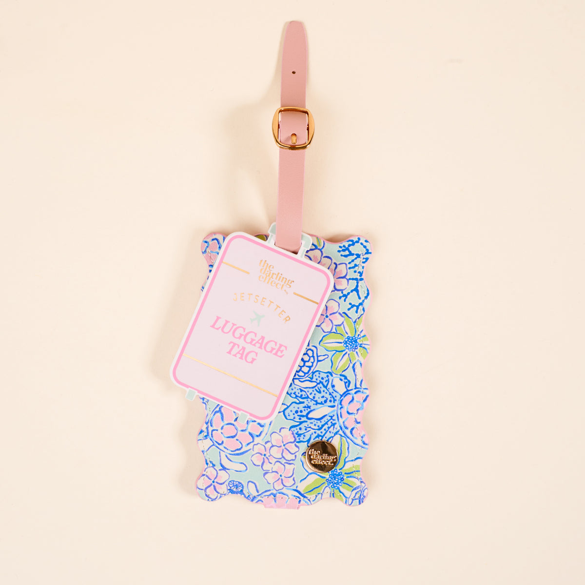 The Darling Effect’s Coastal Cove Jetsetter Luggage Tag features a floral print in pink, blue, and green on a beige background, with a pink strap, gold buckle, privacy flap, and “Jetsetter Luggage Tag” text.