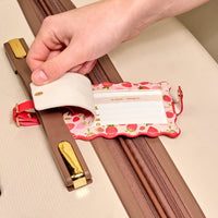 A hand lifts the privacy flap of The Darling Effect’s Strawberry Fields Jetsetter Luggage Tag on a beige suitcase, revealing an information card inside this charming travel accessory.