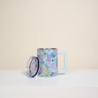 The Darling Effect’s Coastal Cove 14 oz Insulated Mug, featuring a vibrant floral design and a large light blue handle, sits on a white surface with its clear insulated lid beside it against a plain beige background.