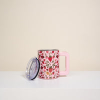 The Darling Effect’s Strawberry Fields 14 oz Insulated Mug has double-wall insulation, a rectangular handle, a white surface with red, pink, and yellow floral patterns, and comes with a transparent lid resting beside it.
