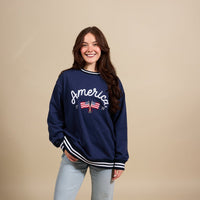 A young woman smiles in The Darling Effect’s America Oversized Crew, a cozy navy blue sweatshirt featuring “America” and an embroidered flag on the front. She poses against a beige background with one hand in her pocket.