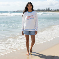 A woman with long dark hair walks barefoot on the sand, wearing The Darling Effect’s Sun Rays, Beach Days, Boat Waves Oversized Crew and denim shorts. Waves roll in behind her beneath a partly cloudy sky.
